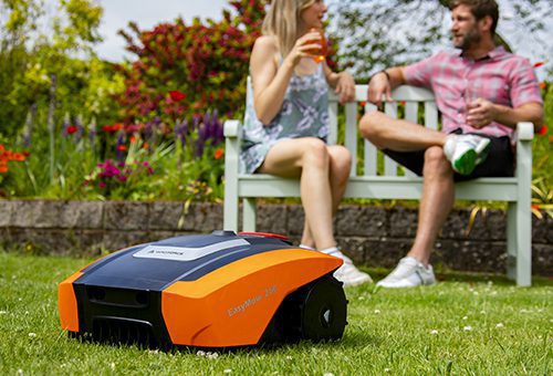 A couple sitting on a bench taking it easy while the robotic lawnmower cuts the grass.