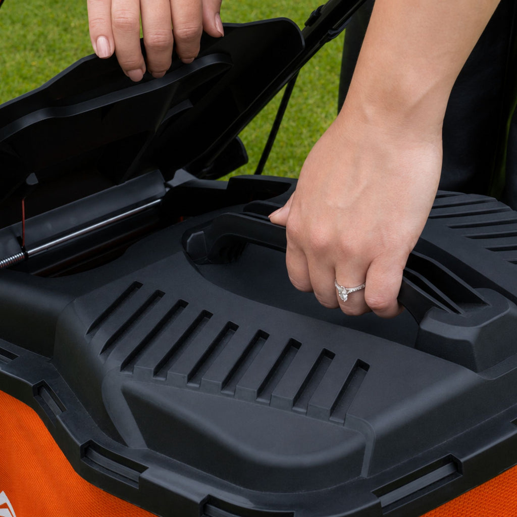 Close-up of a person's hands opening the lid of an orange and black lawn mower with grass visible in the background.