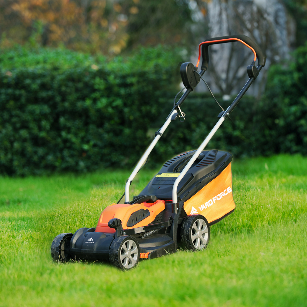 Orange and black Yard Force EM U32 electric lawn mower on a grassy area with green bushes in the background.